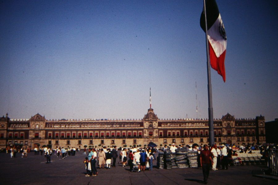 An image of the Mexico City National Palace with a crowd of people standing in front of it and a Mexican flag waving in the foreground.