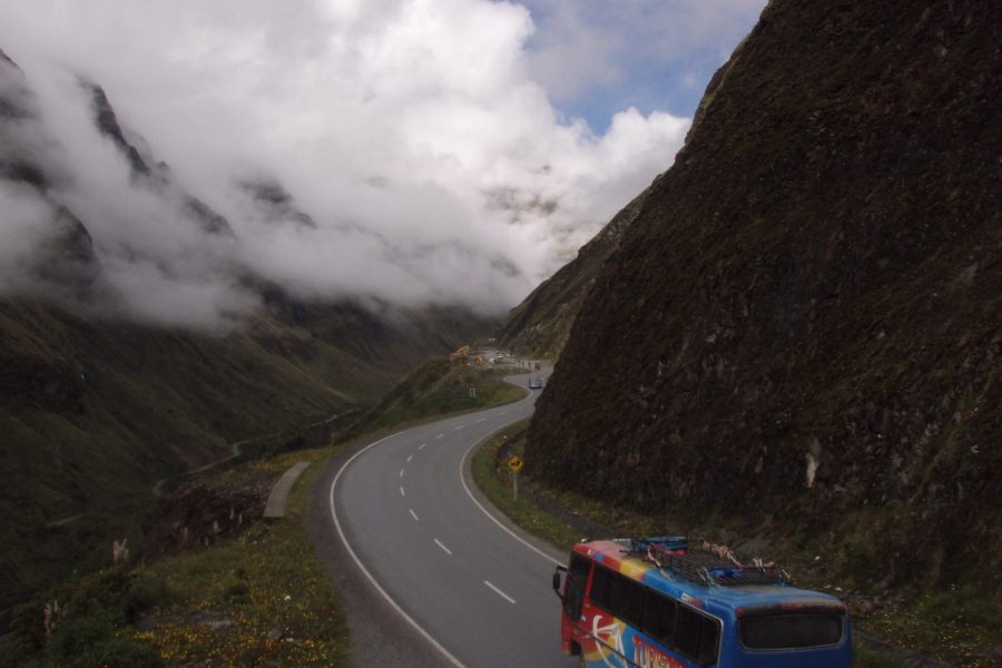 A brightly painted bus travels along a winding mountain road in Bolivia, cutting through a dramatic cloud-covered Andean valley, with steep rocky cliffs on one side.