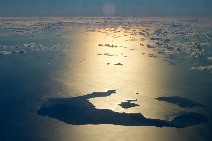 A view of the island of Santorini, Greece, from the air in the late afternoon hours. Golden light shines off the blue Mediterranean sea, while small white clouds spread out over the horizon.