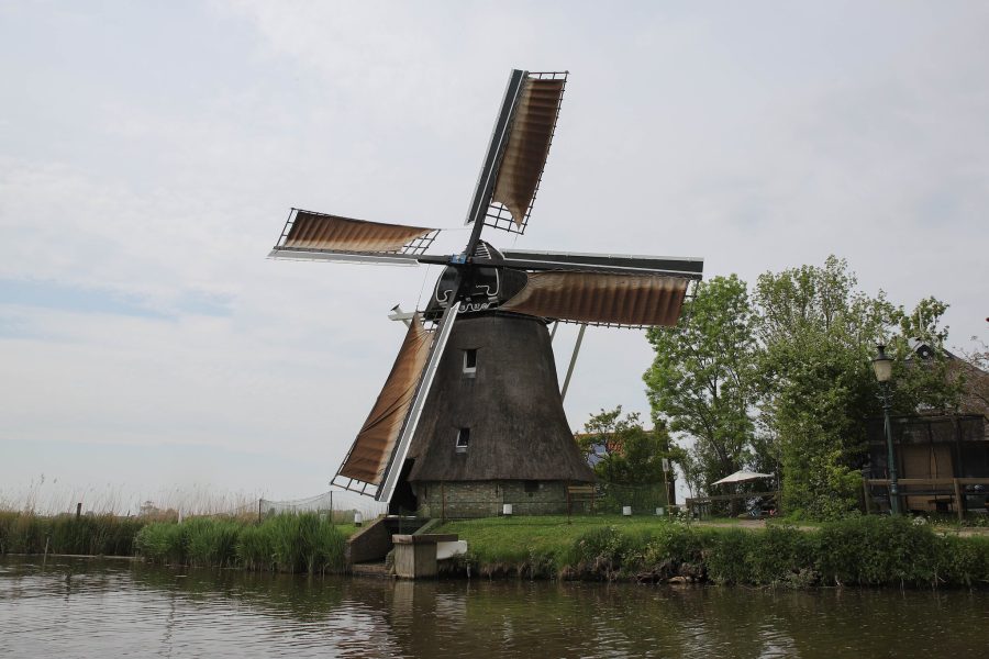 A traditional wood and stone windmill powered mill next to canal, green trees, and wooden structure in the Netherlands.