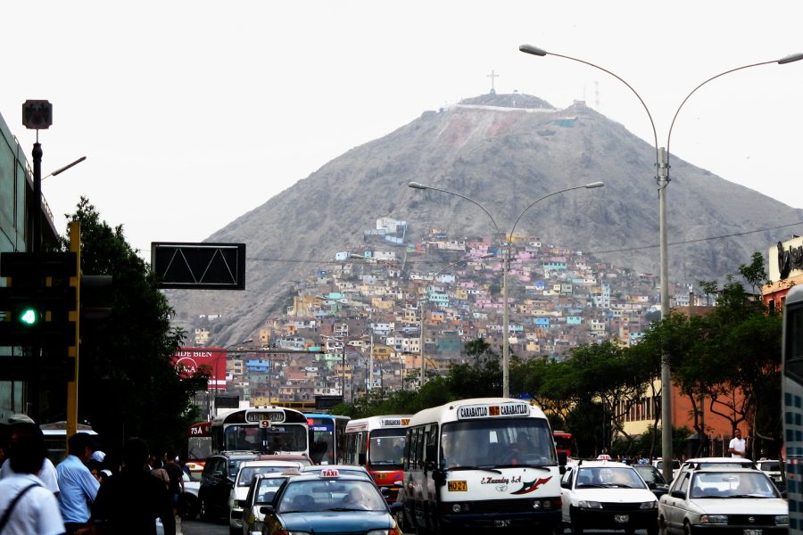 An image of a mountain rising above a busy street in Lima, Peru. The face of the mountain is covered with colorful houses with a large cross on the peak of the mountain.